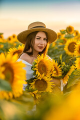 A girl in a hat on a beautiful field of sunflowers against the sky in the evening light of a summer sunset. Sunbeams through the flower field. Natural background.