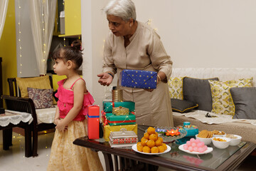 A grandmother offering gift trying to please cute young little girl kid, dressed up in ethnic wear,...