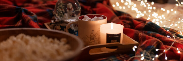 cup of hot chocolate and marshmallows and burning candle on tray in bed and garland glowing lights bokeh at home. taking care of yourself and relaxing on new year and christmas eve dark night. banner