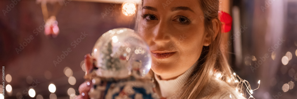 Wall mural beautiful young candid woman holding in hands near face and admire snow globe or dome glass ball and enjoyment an inspiring mood relaxing at home on merry christmas or happy new year eve night. banner