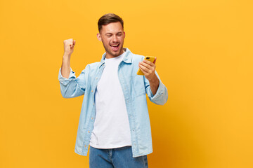 Smiling enjoyed tanned handsome man in blue basic t-shirt raise fist up using phone posing isolated on orange yellow studio background. Copy space Banner Mockup. Online Social media. Lucky win concept