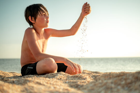A boy sits on the beach on a hot summer day and plays with sand.