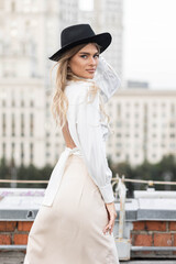 Beautiful attractive young woman with blonde long hair, model on the rooftop. Trendy look, gorgeous outfit, black hat. Cityscape on the background. Unusual modern wedding dress