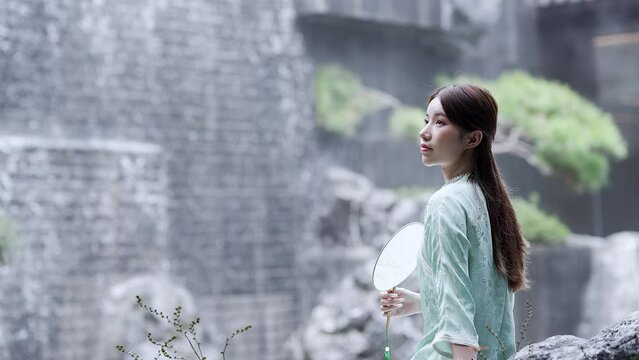 young beautiful chinese woman in qipao relaxing near landscape waterfall wall