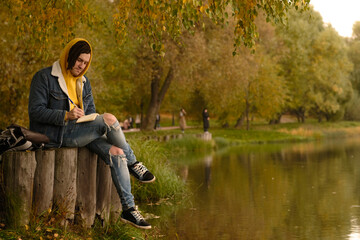 Fototapeta premium Young creative man with dreadlocks drawing, writing in notebook, sitting on stumps near lake in parkland.