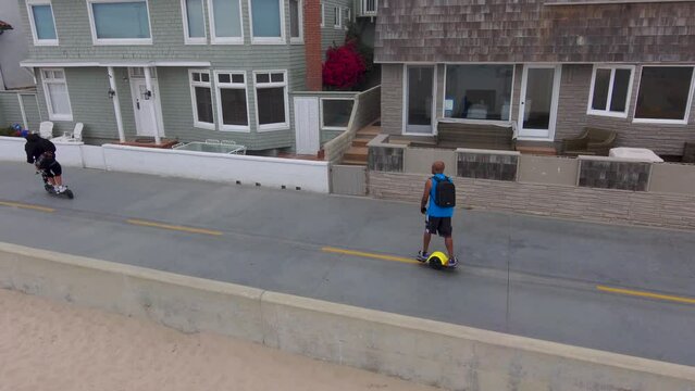 aerial footage of an African American man wearing a backpack riding a Onewheel electric skateboard along a bike path surrounded by buildings and lush green trees and plants in Manhattan Beach