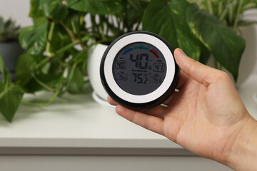 Woman holding digital hygrometer with thermometer near plants indoors, closeup