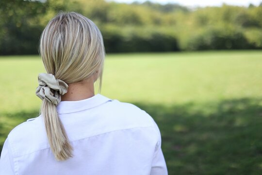 Young Blonde Woman From Behind, Wearing A White Shirt And Scrunchie On Her Ponytail In A Sunny Field