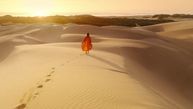 Young beautiful girl in orange linen safari dress walking along sandy dunes in scenic vast desert at ocean coast. 4K Slow Motion aerial back view of traveler hiking barefoot by desert at golden sunset