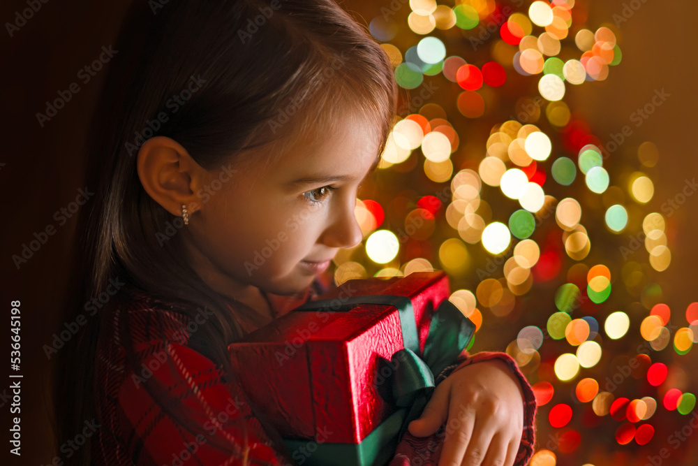 Wall mural a child hugs a gift box in the twilight of christmas night at a christmas tree with colorful lights 