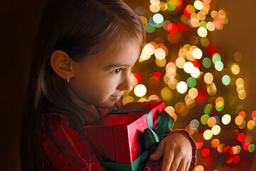 A child hugs a gift box in the twilight of Christmas night at a Christmas tree with colorful lights...