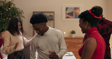 Students dance in the middle of living room. Music is playing loudly and friends are having fun and smiling. Girls and boys move in time. Guy of African appearance in white sweater dances with glass.