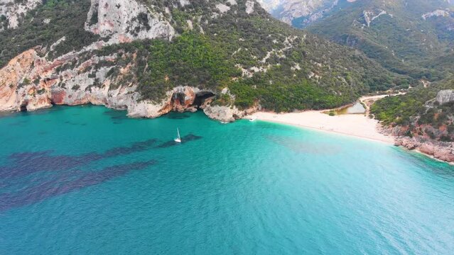 Wundersch&ouml;ne einsame Bucht auf der Insel Sardinien in Italien mit einem Segelschiff auf dem t&uuml;rkisen blauen Meer und Bergen und Strand im Hintergrund 4k