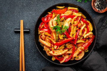 Asian cuisine, stir fry with chicken, red paprika pepper and zucchini in frying pan. Black kitchen table background, top view, copy space