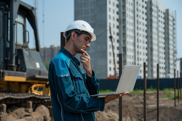 A male builder in a helmet stands against the background of a construction site with a laptop in his hands. 