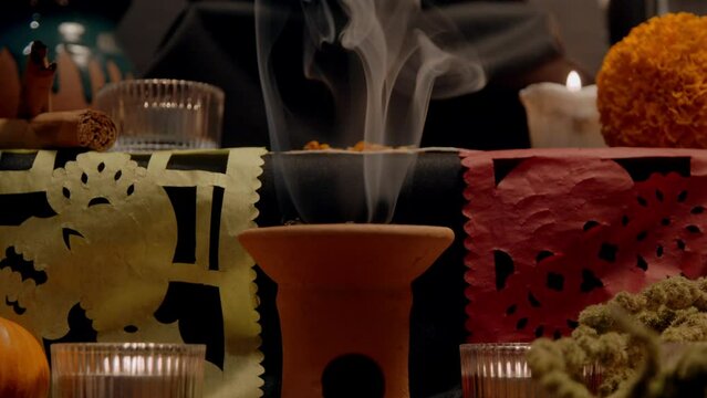 Traditional mexican bread of the dead "pan de muerto" with black ashes in Mexican altar with &ldquo;papel picado&rdquo;, copal, incense burner, cempasuchil flowers. Slow motion, close up, tilt up.