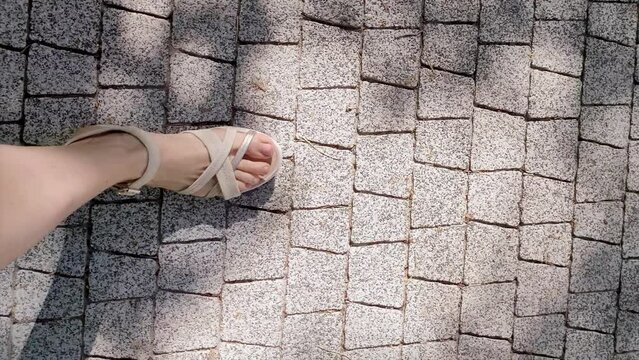 female legs in grey sandals walk along a cobblestone pavemen, first-person view, pov, vertical