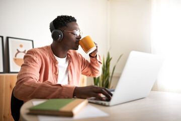 Young African American black man drinking coffee while listening to music and working with laptop. Working from home.