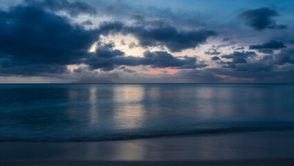 Fototapeta na wymiar Blue hour after sunset on a tropical beach. Clouds over the turquoise ocean. Pink highlights in the sky. Foam waves on the beach. Reflection on water and wet sand. Long exposure. Seychelles. Mahe. 