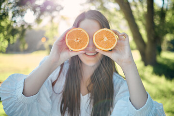 Woman, orange slice and eye pose in outdoor park with natural summer sunshine ray flare. Happy caucasian girl holding healthy, juicy and citrus vitamin fruit for fun outdoor posing moment.
