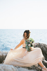 Fototapeta na wymiar Bride with hair fluttering in the wind adjusts her dress sitting on the rocks by the sea