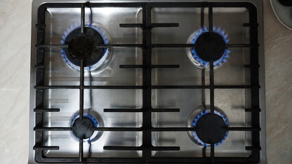 A blue fire is burning on the gas stove. Gas at home. Metal gas stove. Black bars. Light switches of burners. The gas is working. Beige tiles on the walls. Stop the economic crisis. Warmth in house