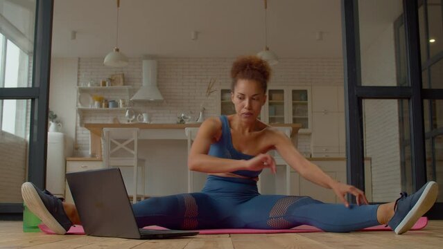 Motivated Active Sporty Fit African Woman Practicing Yoga Class Online On Laptop, Doing Wide Leg Seated Hamstring Stretch Exercise, Increasing Flexibility And Improving Lower Back Muscles Indoors.