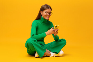 Portrait of woman in green with phone sitting on floor isolated on yellow background