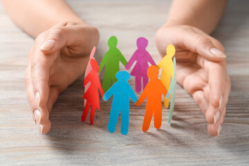 Woman protecting paper human figures on wooden table, closeup. Diversity and inclusion concept - obrazy, fototapety, plakaty