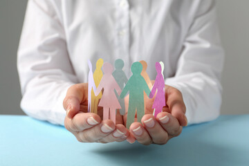 Woman holding paper human figures at light blue table against grey background, closeup. Diversity and inclusion concept