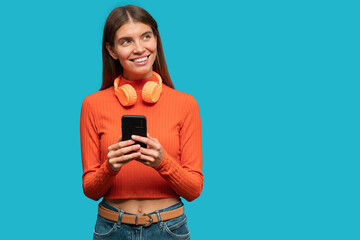 Portrait of dreamy woman holding phone with wireless headphones around neck using music app