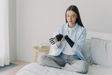 Disabled young girl disassembles bionic prosthetic arm, sitting on bed at home. Medical technologies