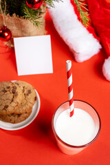 Obraz na płótnie Canvas Milk glass with drinking straw, cookies, empty note, Santas hat on red background. Soft focus.