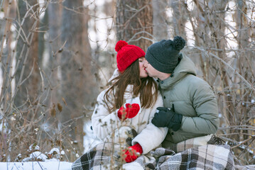 Young couple are sitting hugging and kissing in a snow-covered park, wrapping themselves in a plaid. Man and woman on a picnic in the winter forest.