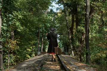 Naklejka na ściany i meble Woman in black coat and hat of 40s style with retro valise in hands walks along rails going through forest. Back view