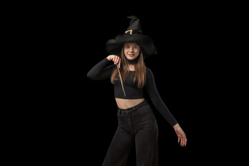 Portrait of young girl looking like witch wearing pointed hat. Teenager with magic wand in hands is isolated on black background.