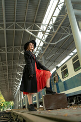 Obraz premium Young woman on platform near train with leather suitcase wearing in red dress, black coat and hat . Vertical frame