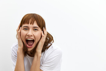 a frightened, shocked, emotional woman screams loudly clutching her face and with very frightened, wide-open eyes stands on a light background in a white T-shirt.