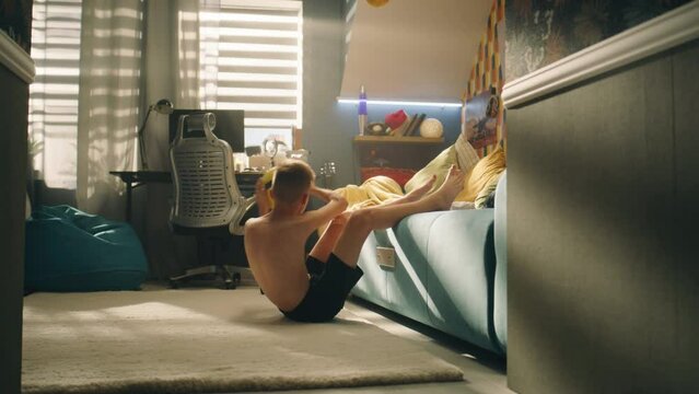 Young boy do abs crunches while holding football in his hands and legs on the couch. Prepares for the football match competition. Soccer, sports, exercising, training and fitness concept.