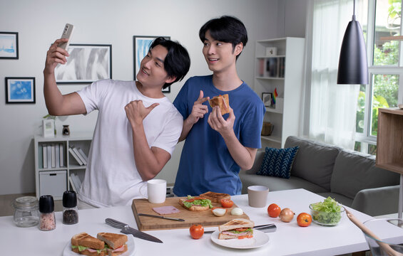 Young Asian gay couple looking happy while cooking and taking selfie. LGBT men couple preparing meal salad in the kitchen at home.