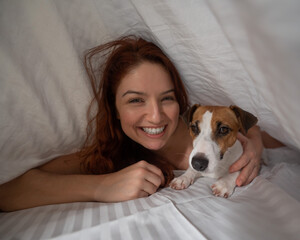 Caucasian red-haired woman sleeps in an embrace with a jack russell terrier dog on a white sheet.