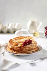 Thin pancakes in milk lie one on top of the other on a plate, currant berries lie on top. Nearby are eggs, flour and honey. Maslenitsa.