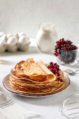 Thin pancakes lie one on top of the other on a plate, currant berries lie on top. Nearby are eggs, flour and honey. Maslenitsa. Traditional cuisine.