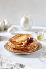 Thin pancakes in milk lie one on top of the other on a plate, currant berries lie on top. Nearby are eggs, flour and honey. Maslenitsa. Traditional cuisine.