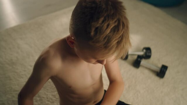 Focused teenage boy with nude upper body exercises. Dumb bells on the carpet. Holds a football preparing before the match. Exercising, fitness, training concept.