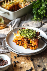 No meat enchiladas casserole dish with black beans, tomato, corn, and sweet potatoes served on a plate on a wooden rustic table with guacamole dip on top