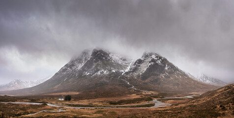Dramatic Winter landscape image of white cottage at foot of Stob Dearg Buachaille Etive Mor peak in Scottish Highlands
