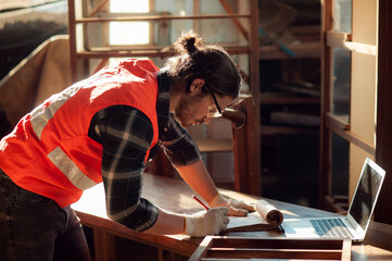 Young and handsome caucasian carpenter in the carpentry shop is working and checking customer's order in laptop to repair a furniture. small business and freelance craftsman.