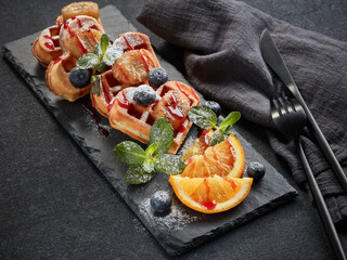 Delicious waffles heart shape with caramelized banana and berries on serving board on grey background. Dessert. Serving food