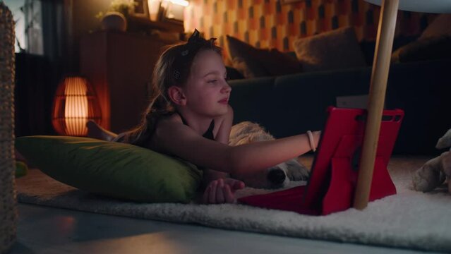 Girl lying on mild carpet with dog, choosing movies to watch, touching digital tablet computer, smiling and chilling, spending leisure time at home. Golden retriever.